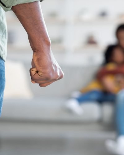 Domestic Violence. Unrecognizable African American Man Threatening Wife And Daughter With His Fist, Scared Mother Embracing Little Girl While Sitting Together On Couch, Selective Focus On Male Hand